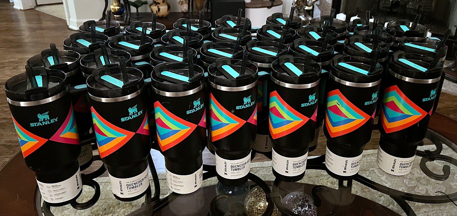 Rows of new black Stanley Quencher tumblers with a bright multicolored geometric design and white product labels, lined up on a glass table
